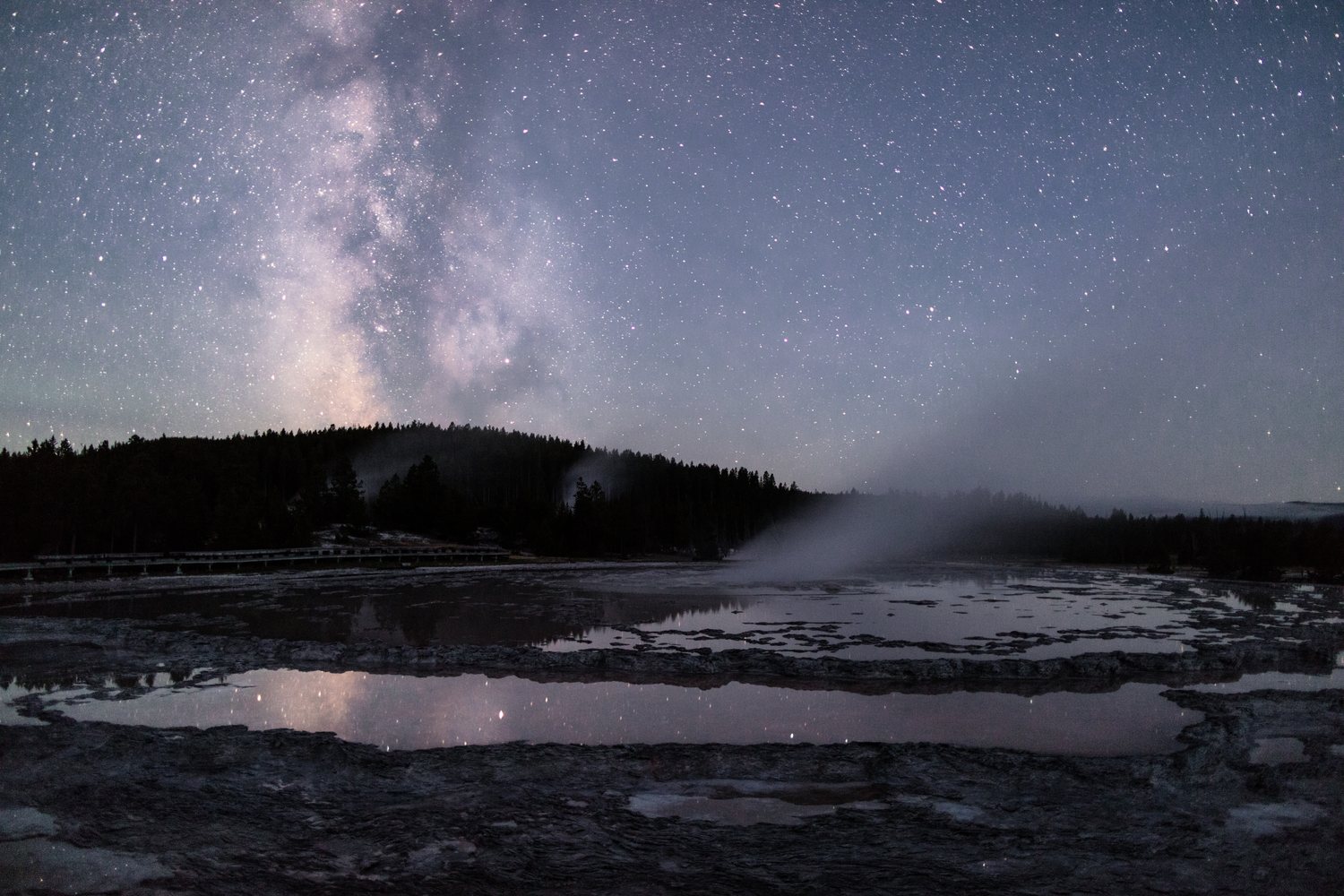 The Milky Way reflected in the still pool of Great Fountain Geyser with a plume of steam against the stars.